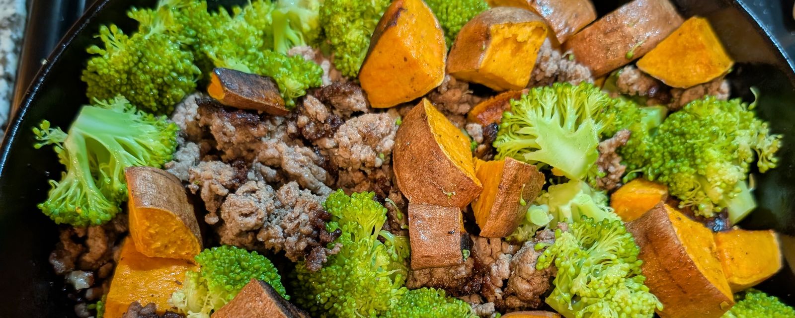 A cast-iron skillet of ground beef, roasted sweet potato, and broccoli on the stove