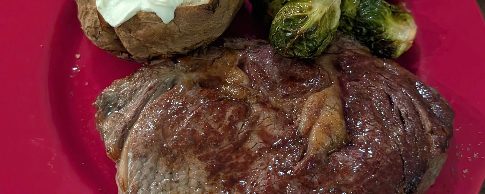 A home-cooked ribeye with a baked potato and roasted Brussels sprouts on a red plate