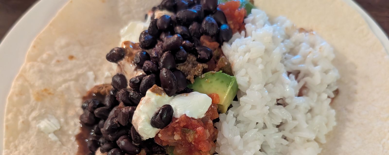 A tortilla topped with black beans, rice, avocado, salsa, and crumbled cheese on a plate